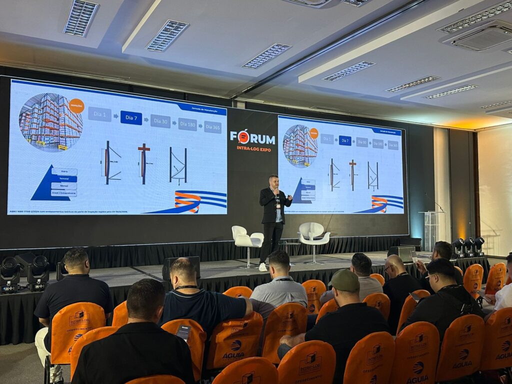 A imagem mostra o palco do espaço de palestras na feira e telas sendo apresentadas por consultor de vendas da Bertolini Sistemas de Armazenagem na feira INTRA-LOG 2025. As informações nas telas são sobre a jornada de manutenção em sistemas de armazenagem. Várias pessoas assistem à apresentação, em ambiente de fundo escuro.
